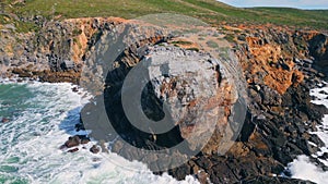 Rugged coastal cliff washing by stormy foamy ocean sunny day. Drone rough sea