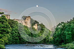 A ruin of a castle protruding through a forest next to a river with a moon