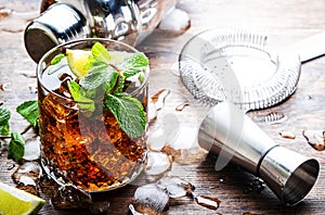 Rum cola cocktail drink with strong alcohol, mint, ice and lime in glass. Wooden background, hard light