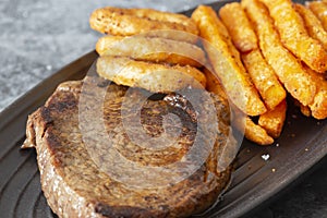 Rump steak dinner, with sweet potato fries and onion rings, on a black oval plate.