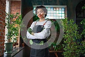 Running of own business. African woman florist wearing apron in botanical store with green plants. Happy small business