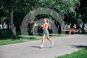 running smiling young sporty woman in park in sportswear on background of green trees on grey tiles in sunny windy