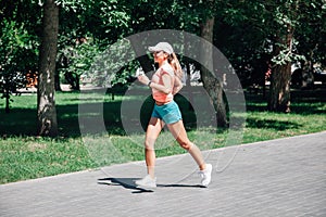 running smiling young sporty woman in park in sportswear on background of green trees on grey tiles in sunny windy