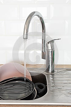 Running tap water in metal kitchen sink with dirty dishes. Needing washing-up. Cleaning chores. Professional service cleaning