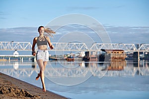 Running woman in the sand at sunrise. Morning jogging barefoot on the beach or coast of river on urban city background