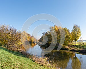 Rural autumnal river scene with blue sky, colorful trees reflecting on the water, a bench and