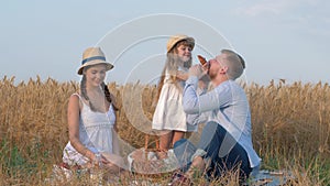 Rural family portrait, little happy kid girl eats yummy sweet bun and treats her parents at village picnic in reaped