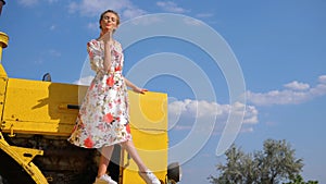 Rural girl in dress gives air kiss on bonnet of combine harvester on sky background