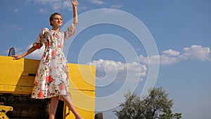 Rural girl into dress Waving of Hand standing on bonnet of tractor at nature on background of sky