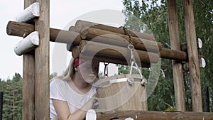 Rural girl looking in wooden bucket on water well in summer village. Young country woman checking water in bucket on