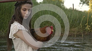 Rural girl in white dress in cuddles with rooster on the river, relax rutal life