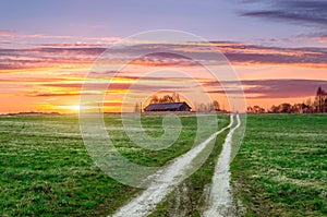 Rural landscape, a road in the field rises to a hill with a standing barn during the evening summer sunset.