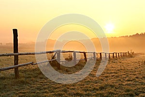 rural landscape at sunrise with wooden fence in the field in misty weather wooden fence in the field lit by rising sun in foggy