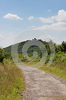 Rural old asphalt road through forest turning left. Historical castle ruin on hill in centre. Summer sunny day in Slovak nature