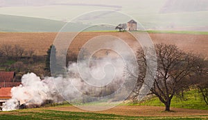 Rural Pastoral Spring Landscape With Old Windmill Without Blades And Plowed Land On Background Of The Village And Thick White Smok