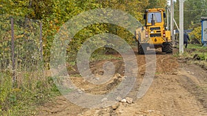 Rural road construction with grader in autumn daytime. The grader repairs the dirt road in the village.