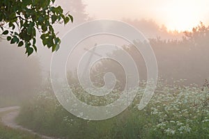 Rural road and old leaning cross between tall grass and trees in fog at sunrise