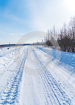 Rural road in winter covered with rolled snow on the background of snow-covered fields and forests with a clear sky