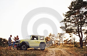 Rural scene. Group of cheerful friends have nice weekend at sunny day near theirs green car outdoors