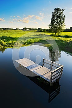 Rural summer landscape with wooden platform in river and green meadows with growing willow tree.