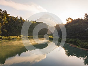 Rural view of lake surrounded by dense of forest with mountain background and blue sky in sunrise time