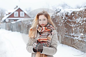 Rural young stylish woman in a winter gray coat in a vintage stylish scarf in warm woolen mittens is standing on a snowy road