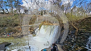 rushing water flowing over a waterfall surrounded by rocks, green trees and plants, people, blue sky and clouds and homes