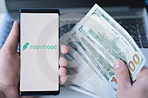 Russia Moscow 02.02.21 Mobile phone with logo of broker Robinhood, money, dollars in hand.Financial trading company, investment in