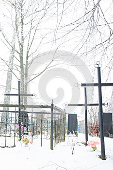 Russian village cemetery: Multiple types of fencing, blue crosses, tombstones, monuments, flowers, trees and shrubs, on a cold day