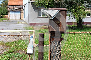 Rusted broken mailbox with faded newspaper mounted on wooden pole behind wire fence with abandoned family house in background