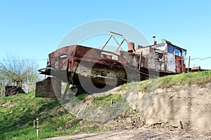 Rusted metal river barge left unused on edge of river bank surrounded with grass and dry soil on clear blue sky background