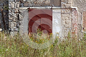 Rusted metal sliding warehouse doors mounted on old dilapidated wall with tall grass and small flowers in front at abandoned