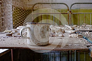 A rusted mug on a dilapidated table in an abandoned room. A white metal mug on a bed in an abandoned building in Chernobyl