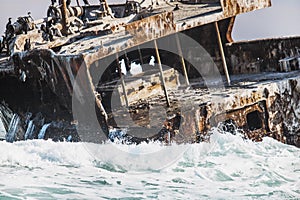 Rusted shipwreck on the shore with crashing waves and Cape Cormorant birds.