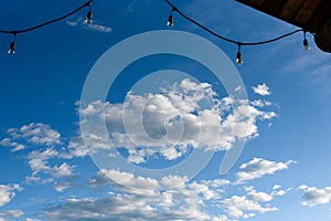 Rustic background of outdoor wood ceiling and string of lights framing a blue sky with white clouds