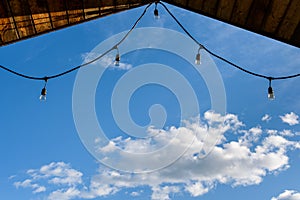 Rustic background of outdoor wood ceiling and string of lights framing a blue sky with white clouds