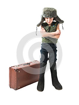 Rustic boy in a fur hat and boots standing near an old suitcase with his hands crossed