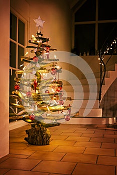 Rustic Christmas tree made of raw wood branches decorated with glowing fairy lights and red balls in a stairwell at night