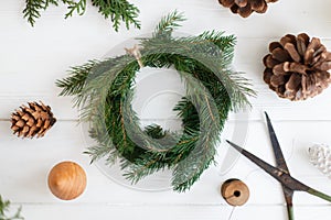 Rustic christmas wreath with pine cones, scissors and ornaments on white wooden table, flat lay