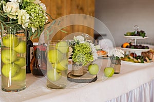 Rustic flower arrangement with white flowers and greenery in a glass vase with water and apples at a wedding banquet. Table set fo