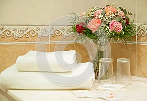 Rustic hotel bathroom necessities featuring a set of white towels, basic glasses, soap and a roses bouquet