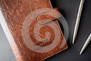 Rustic leathered Employee handbook on office desk with engraved text next to two steel pens on black background with side light.