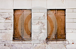 Rustic old grungy and weathered brown wooden closed windows shutters with peeling paint on a white cracked wall