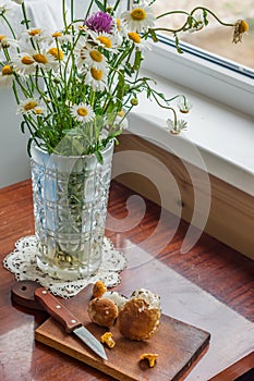 Rustic scene - old-fashionned glass vase with bouquet of chamomiles and wooden desk with mashrooms and knife on window background photo