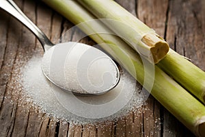 Rustic setup with granulated sugar spoon and sugarcane on wooden surface.