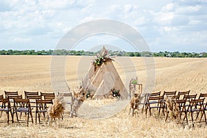A rustic-style wedding ceremony venue nestled in a rural setting. A triangular wooden arch, adorned with flowers, graces the