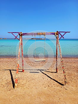 Rustic swing on the beach sand, sea and turquoise water under blue sky of the Dahab desert. Wooden and rope swing. Games and