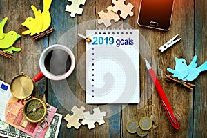Rustic table with spiral notebook with `2019 goals`, cup of hot coffee, smartphone, money, compass, pen and other objects for deco