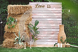 Rustic wedding photo zone decorated with wooden background, hay, wicker pots with plants, lamps and candles. Copy, empty