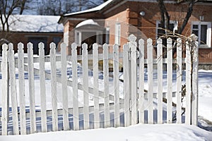 Rustic winter landscape with a wooden fence and a snow-covered little house behind it. A sunny day with a blue clear sky